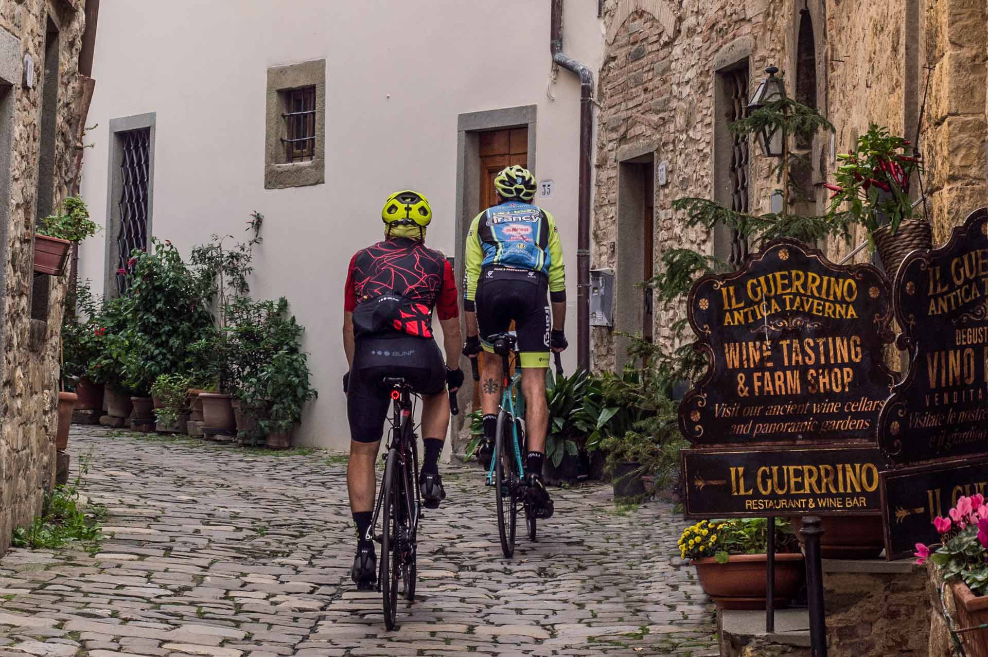 Due ciclisti percorrono una strada acciottolata tra le vie storiche di Castellina in Chianti, vicino a un’enoteca con insegna "Il Guerrino"